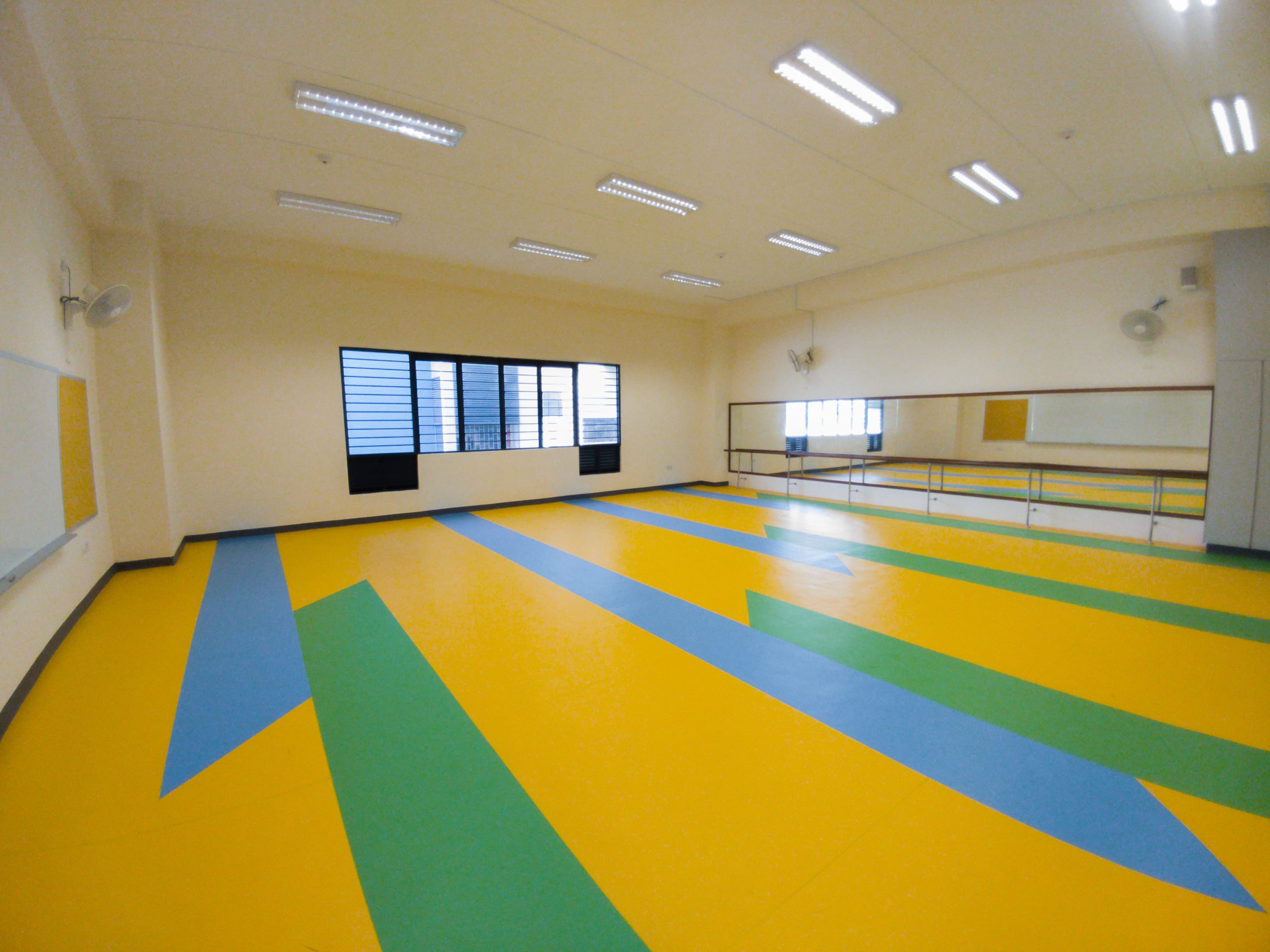 Empty dance studio with yellow, green, and blue patterned floor, wall mirrors, ballet barre, and fluorescent ceiling lights.